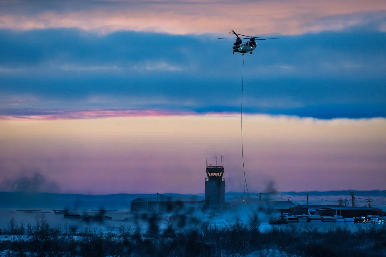 A helicopter carries a suspended load above a snowy settlement at dusk.