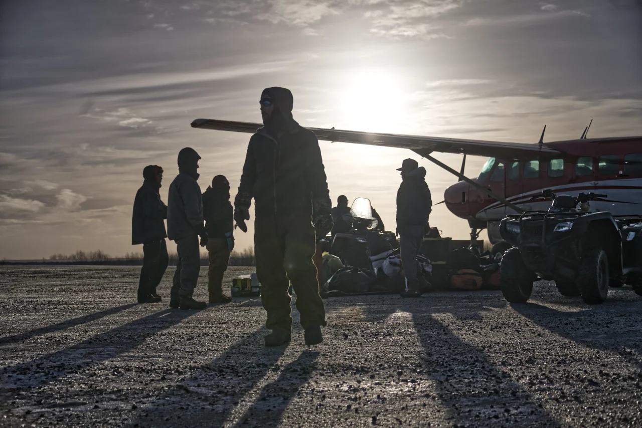 People and equipment beside a small aircraft on an airstrip, silhouetted by low sunlight.