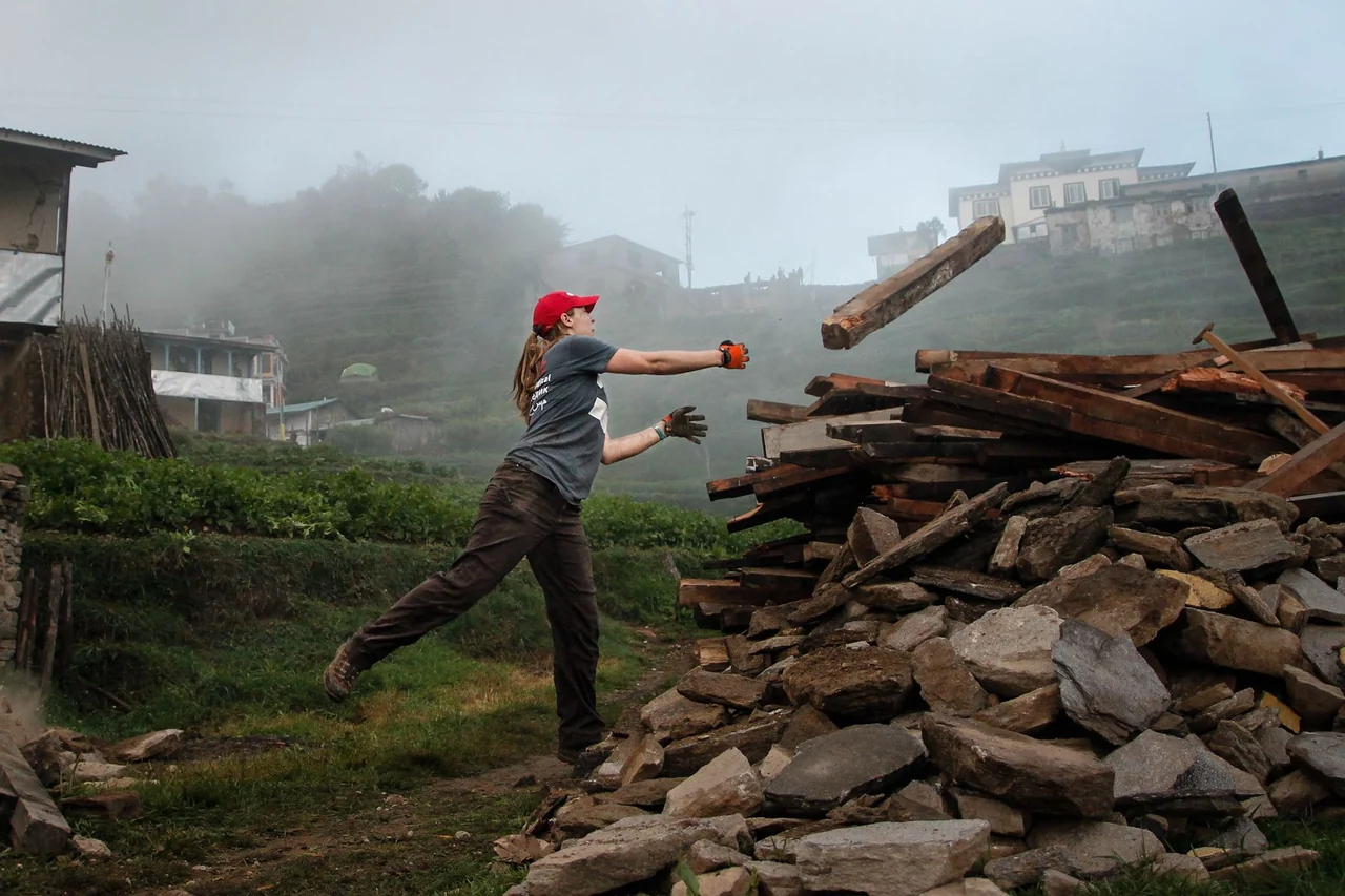 A volunteer in a red cap throws a timber beam onto a pile of debris in a misty hillside settlement.