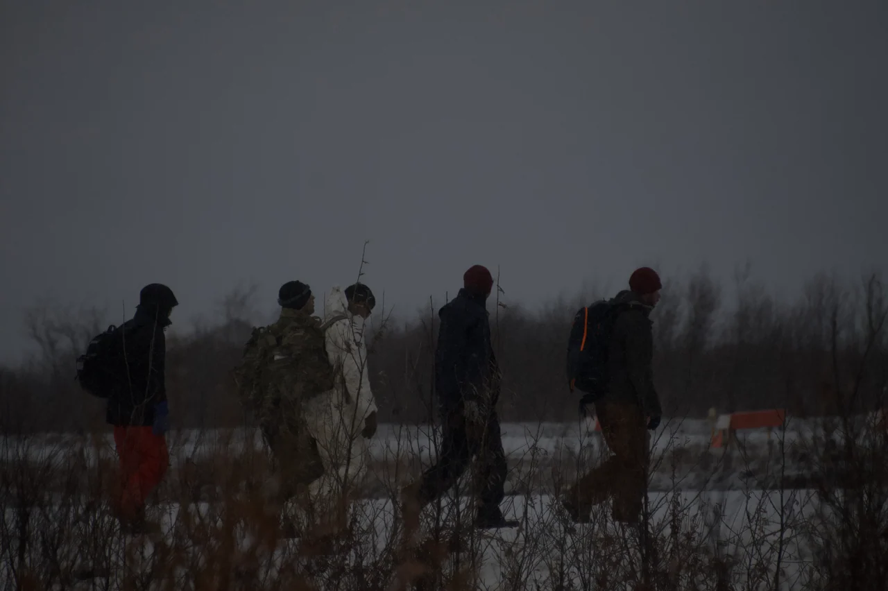 A line of people walks through low vegetation in dim winter light.