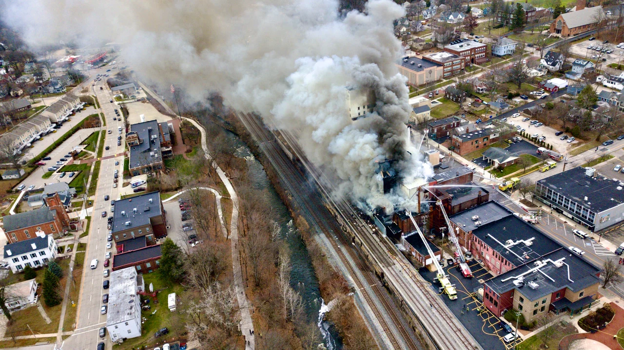 Smoke billows from a large building beside railway tracks, seen from the air.