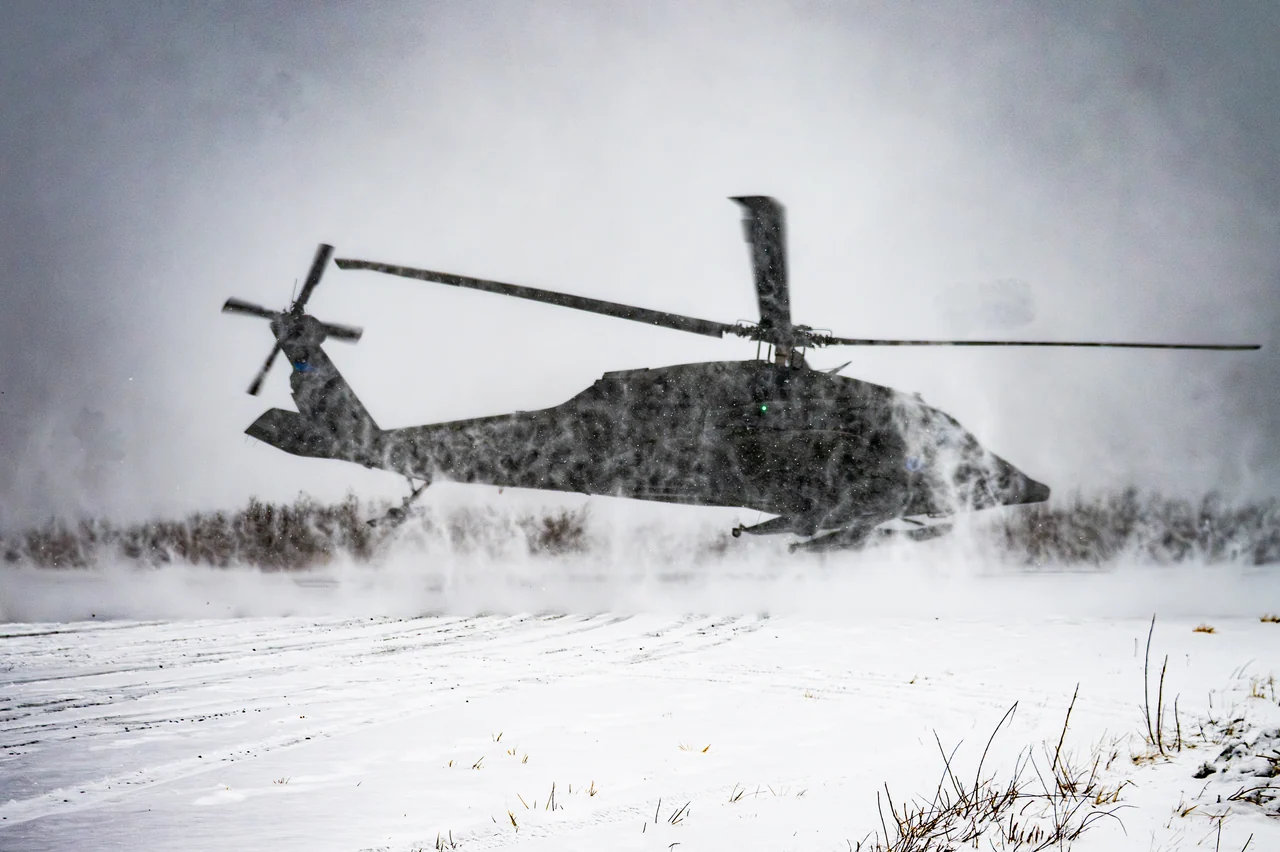 A helicopter sits in blowing snow on a white field.