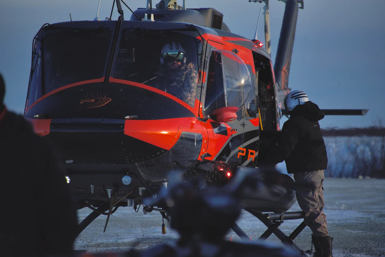 A bundled figure stands beside a red helicopter in the snow.