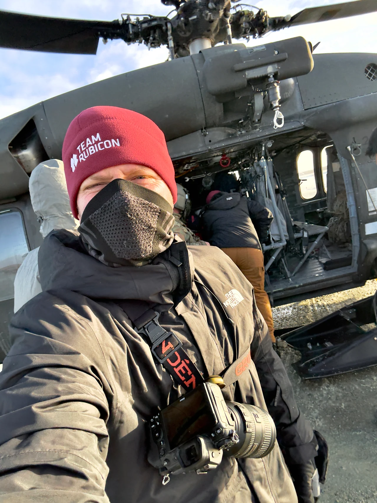 Josh Walker stands beside a helicopter wearing winter clothing and a Team Rubicon hat.
