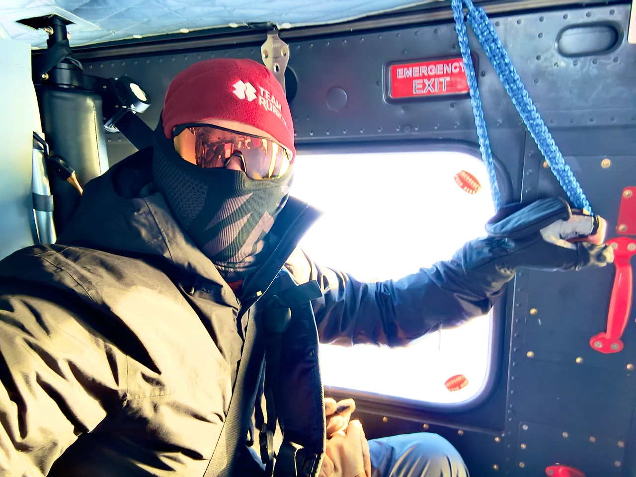 Josh Walker sits in a helicopter cabin wearing a red hat, reflective glasses and a face covering.