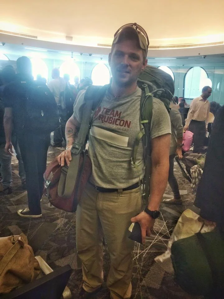 Josh Walker stands indoors wearing a large backpack and outdoor clothing.