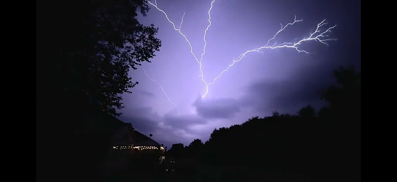 Branches of lightning illuminate a purple night sky above silhouetted trees.