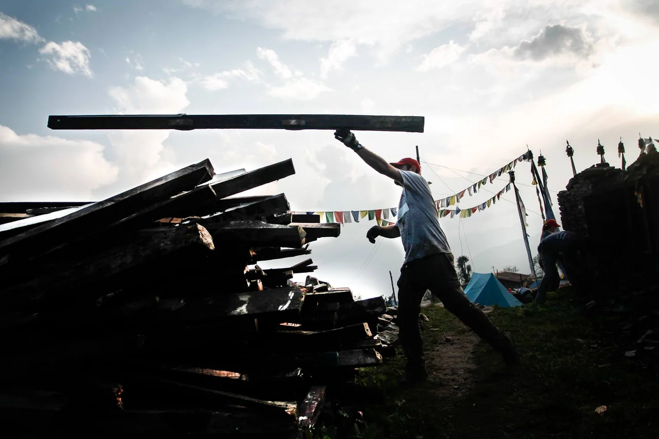 A worker carries a long beam above a pile of timber, silhouetted against the sky.