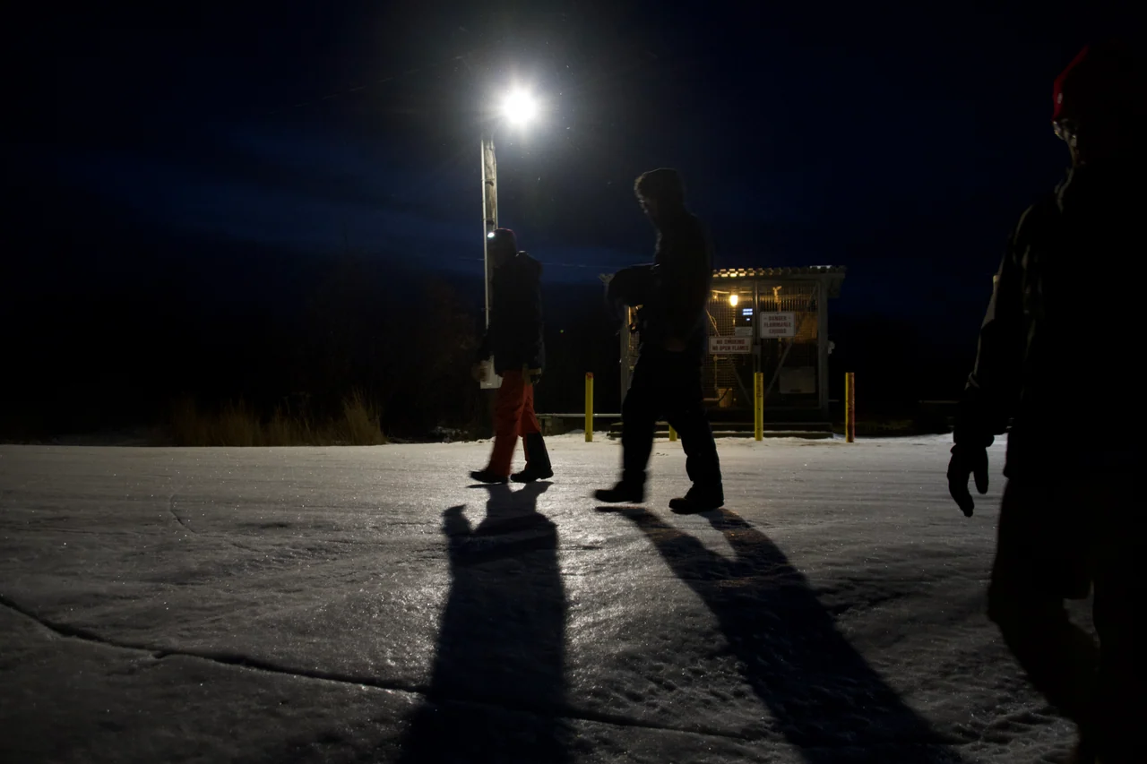 People cross an icy open area beneath a bright light at night.