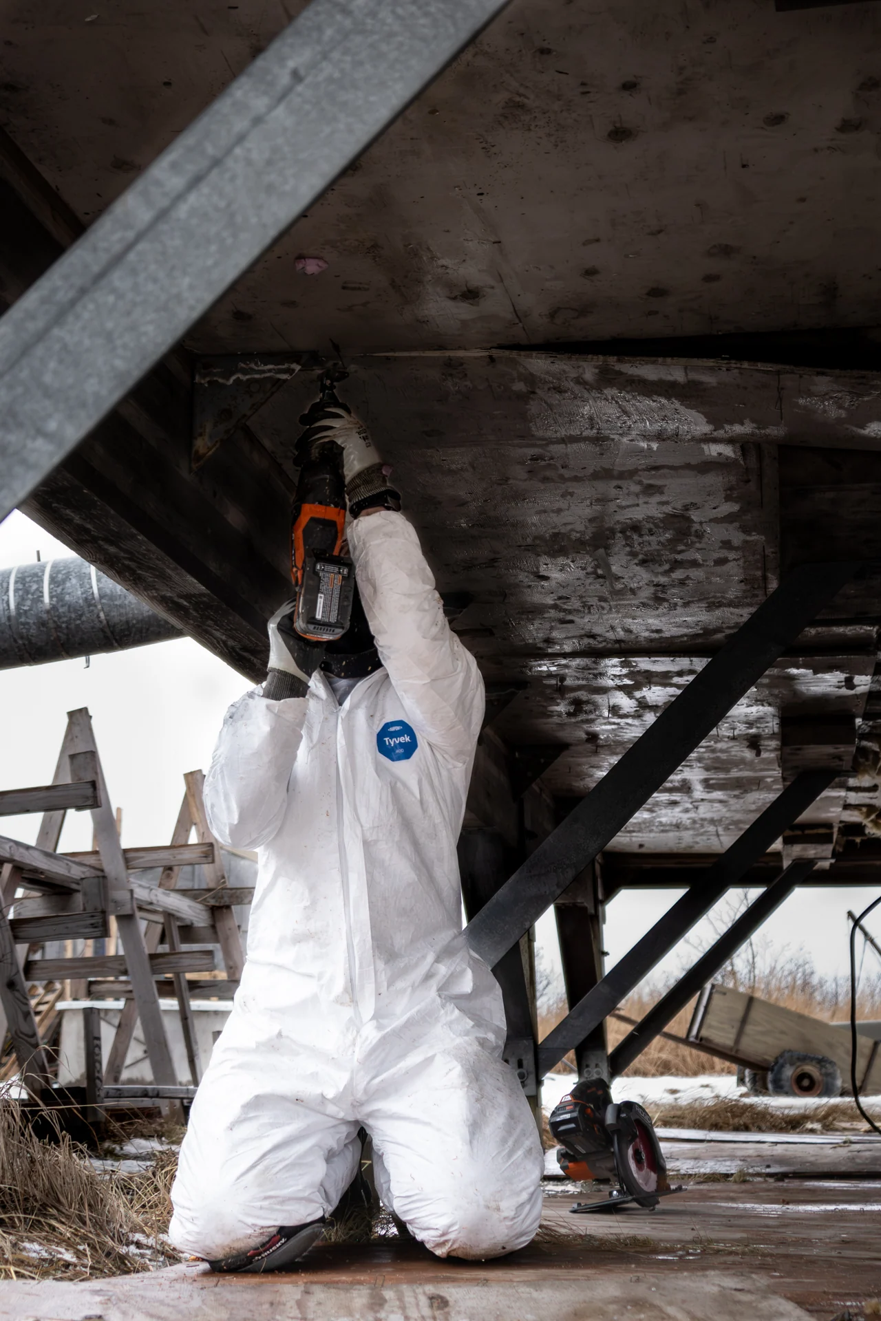 A volunteer in white protective clothing kneels beneath a raised building.