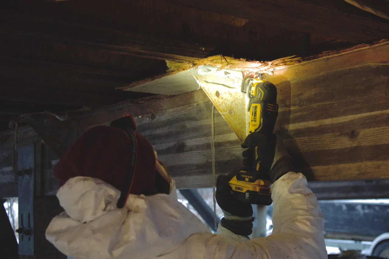 A volunteer uses a power tool on timber beneath a building, illuminated by a work light.