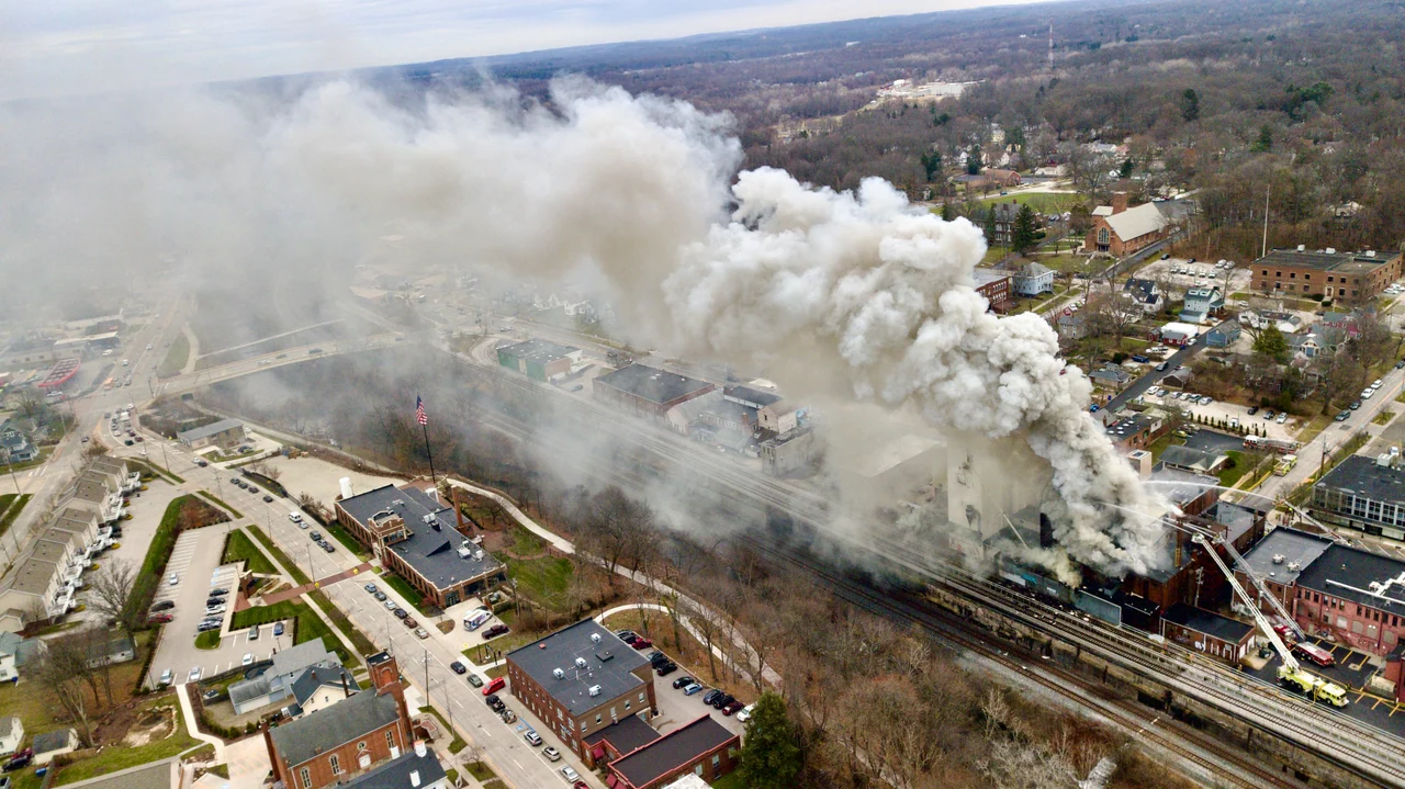 An aerial view shows a broad smoke plume rising near railway tracks and buildings.