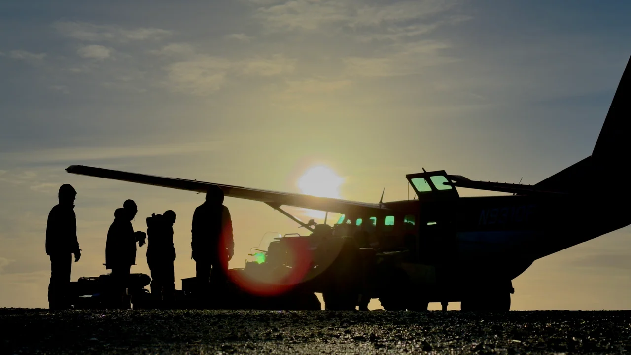 People stand in silhouette beside a small aircraft with the sun low behind its wing.