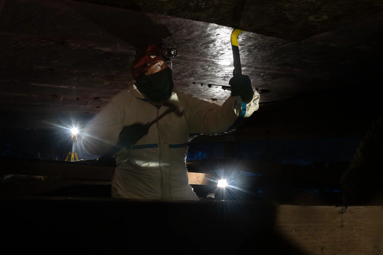 A masked volunteer works with a tool beneath a building, lit by headlamps.
