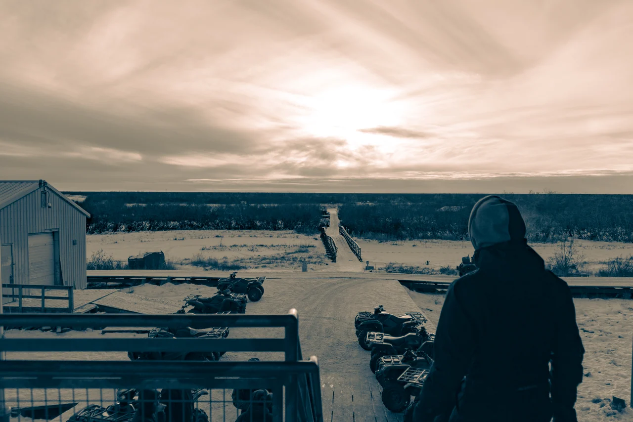 A person stands on a porch overlooking a snowy settlement beneath a low sun.