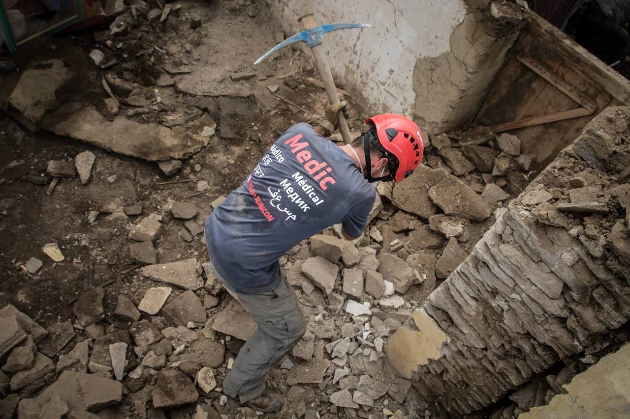 A volunteer wearing a red helmet works among collapsed stone walls, seen from above.