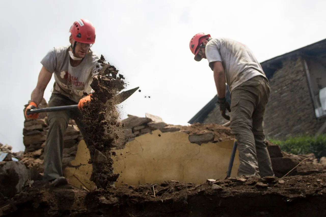 Two helmeted volunteers use tools to clear damaged building materials.